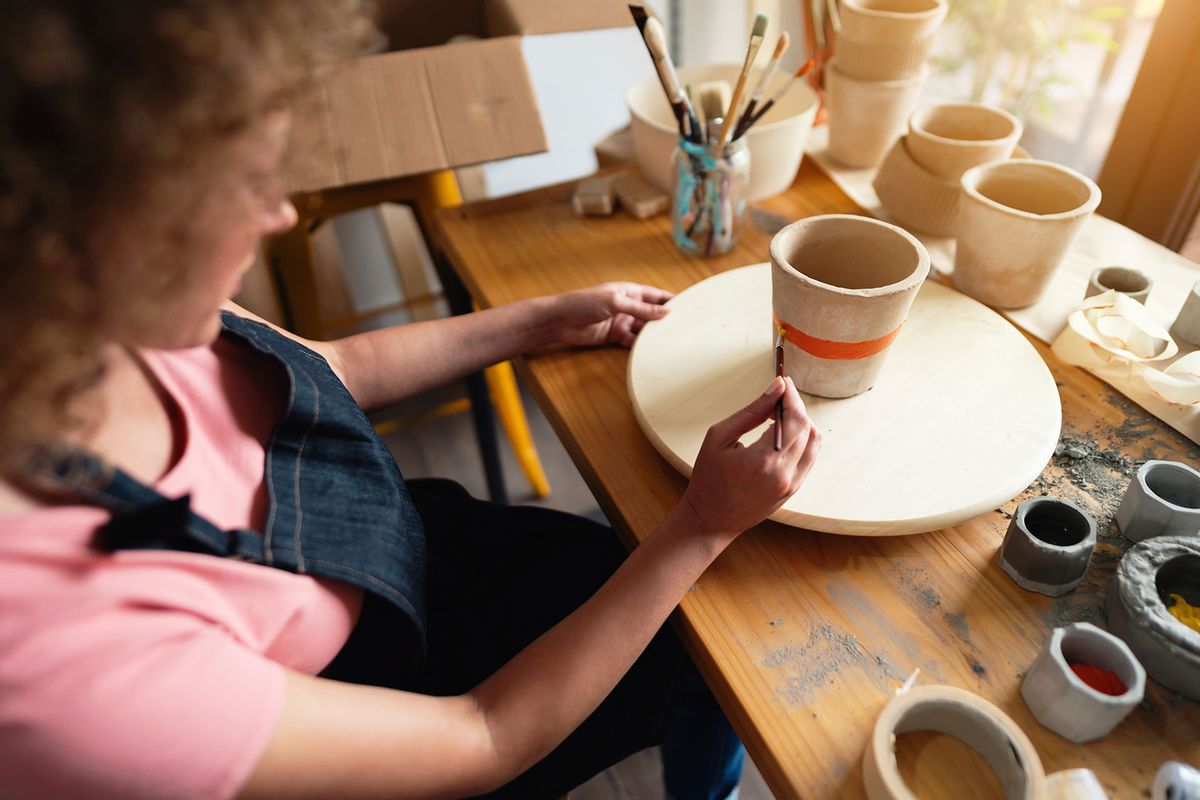 Craftswoman painting ceramic pot in her workshop (Getty Images/SrdjanPav)