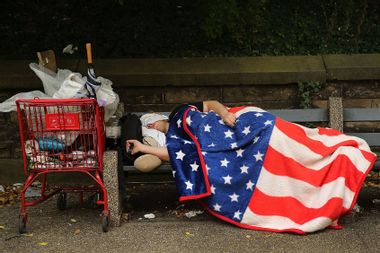 A homeless man sleeps under an American Flag blanket on a park bench.