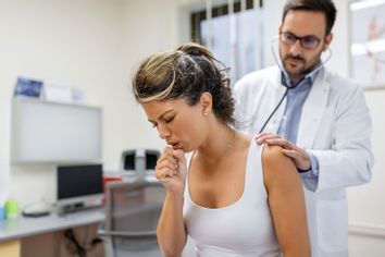 Young female patient in the clinic suffering a coughing fit while the doctor listens to the her lungs with a stethoscope.